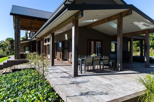 A contemporary multi-level home with a large covered outdoor dining and entertaining terrace. The structure features dark metal pitched roofing, timber posts and beams, broad roof overhangs, dark rendered walls, extensive glazing, and a generous paved deck overlooking landscaped gardens.