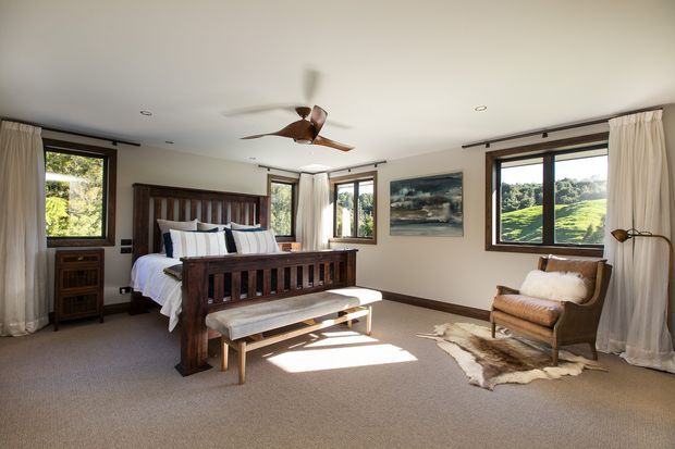 Spacious, warmly lit bedroom with a dark timber four-poster-style bed and matching built-in bedside storage, upholstered bench, neutral carpet, cream curtains, and a leather lounge chair. Large timber-framed windows overlook a green rural landscape, while a ceiling fan and recessed lighting complete the relaxed traditional interior.