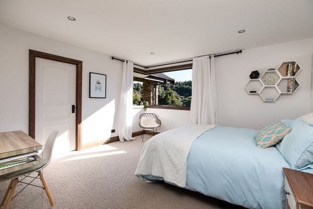 Bright, contemporary bedroom with a large bed dressed in pale blue and white bedding, beige carpet, white walls, recessed ceiling lights, and a dark timber-trimmed internal door. Floor-to-ceiling curtains frame a corner window overlooking greenery, while geometric hexagonal wall shelving provides a decorative feature. A small desk and chair sit near the door.