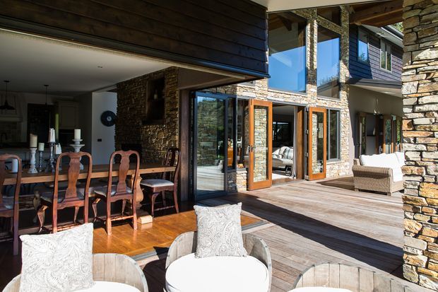 A sunlit multi-level residential courtyard or terrace framed by natural stacked-stone walls and dark timber cladding. Large timber-framed glazed doors open from an interior dining area onto a broad timber deck, with outdoor lounge seating in the foreground. The architecture features exposed timber structure, substantial stone columns and walls, and expansive glazing connecting indoor and outdoor spaces.