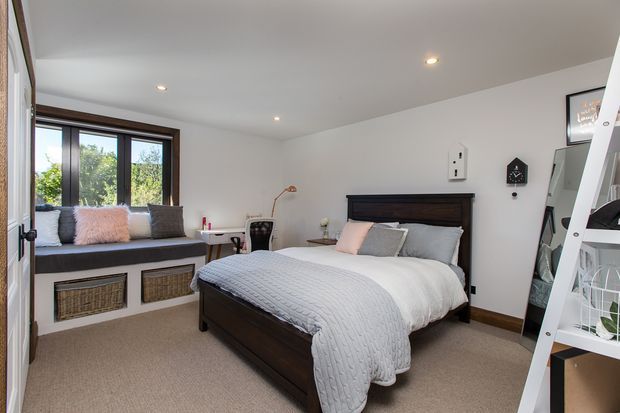 Bright, neatly styled bedroom with a dark timber bed and matching built-in window seat, layered grey and blush bedding, beige carpet, white walls, and a large dark-framed window. A compact desk and chair sit beside the window, while a leaning mirror and white ladder-style shelving occupy the right side.