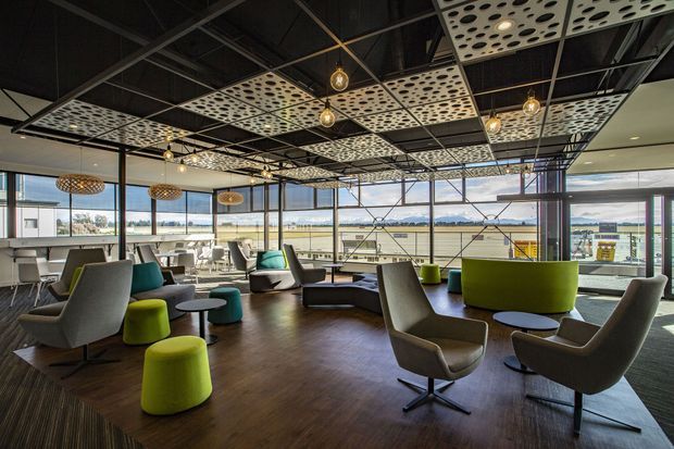 A spacious contemporary commercial lounge or breakout area with floor-to-ceiling glazing, an open-plan seating arrangement, and views across an industrial or urban landscape. The room features dark exposed ceiling services and structural framing, suspended perforated acoustic ceiling panels, pendant lighting, timber-look flooring, and a mix of lounge chairs, low tables, and bright lime-green upholstered seating.