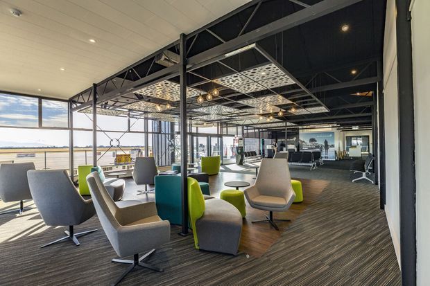 A spacious contemporary commercial lounge or reception area with abundant glazing, views across an open landscape, exposed dark steel roof structure, patterned suspended ceiling panels, pendant and recessed lighting, and varied upholstered lounge seating arranged over a dark textured carpet. The space has a strong industrial-modern character softened by bright green, teal, grey, and beige furniture.