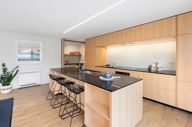 A bright, contemporary kitchen featuring a large light timber island with open shelving, a dark stone benchtop, integrated black appliances, and full-height timber cabinetry. Four black wire-frame bar stools line the island. A recessed illuminated splashback and linear ceiling lighting add a clean, refined finish, while timber flooring and indoor greenery bring warmth.