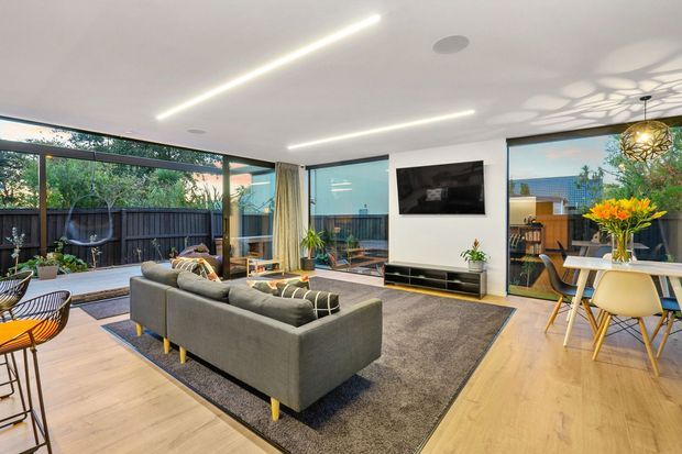 Bright open-plan living and dining area with floor-to-ceiling glazing opening to a landscaped outdoor space. The room features light timber flooring, a large grey sectional sofa, a dark area rug, white walls and ceiling, a wall-mounted television with low built-in cabinetry, and a contemporary dining setting with pendant lighting. Linear recessed ceiling lights and extensive glazing create an airy, modern interior.