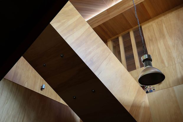 Upward-looking interior view emphasizing warm timber wall and ceiling linings, a prominent angular timber-clad structural or stair element, exposed framing, and a hanging industrial-style pendant light. The composition highlights dramatic geometry and natural wood surfaces in a compact, vertically articulated space.