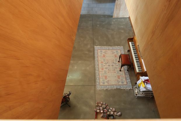 A high-angle view down a narrow, tall interior passage framed by warm timber-clad walls. The polished concrete floor contains a small patterned rug, an upright piano with a wooden finish, a red chair, and a few decorative objects. Exposed grey masonry and timber elements are visible at the far end, creating a warm industrial character.