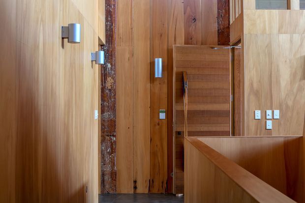 Interior view of a warm timber-lined hallway or landing, featuring extensive vertical and wall-to-wall wood panelling, a built-in timber storage cupboard, modern cylindrical wall sconces, and a timber balustrade in the foreground. A dark textured structural or feature column contrasts with the lighter wood surfaces.