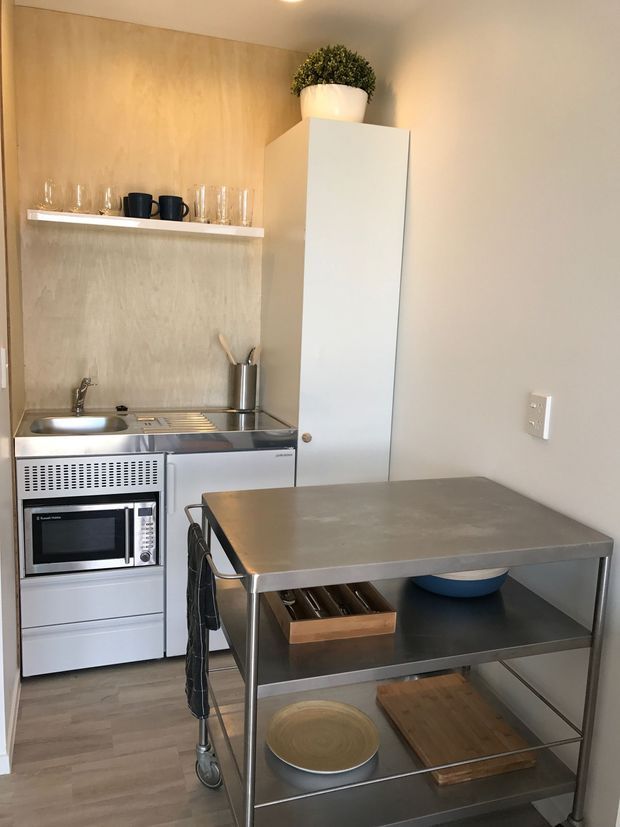 Compact, minimalist kitchenette with stainless-steel appliances and work surfaces, a timber-look wall panel, open shelving with glassware, white cabinetry, and a freestanding stainless-steel worktable with storage shelves.
