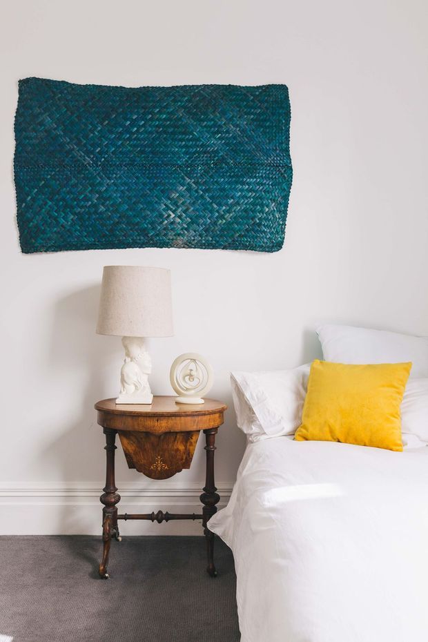 A calm bedroom corner featuring a white bed with crisp bedding, a bright yellow accent cushion, and an ornate dark timber bedside table with turned legs and a sculptural central apron. A vintage-style table lamp and decorative object sit on the table, while a large teal woven textile artwork provides a strong wall accent. The room has white walls with simple lower trim and a grey carpeted floor.