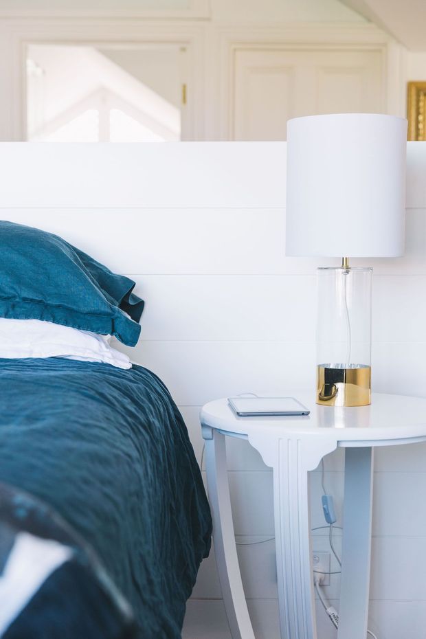 Close-up bedroom interior featuring a white paneled headboard and bedside table, deep teal-blue bedding, and a white table lamp with a clear glass and gold-accented base. The room has bright, refined coastal-inspired styling with traditional wall and door detailing in the background.