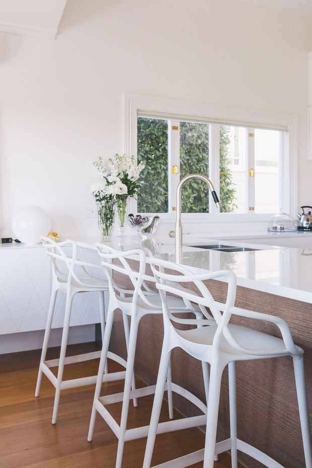 Bright, airy contemporary kitchen photographed from the dining side, featuring a large white island with a dark stone benchtop, integrated sink and tall curved chrome tap. Several white bar stools line the island, while white cabinetry, a timber floor, large framed windows, and a simple floral arrangement complete the clean interior.