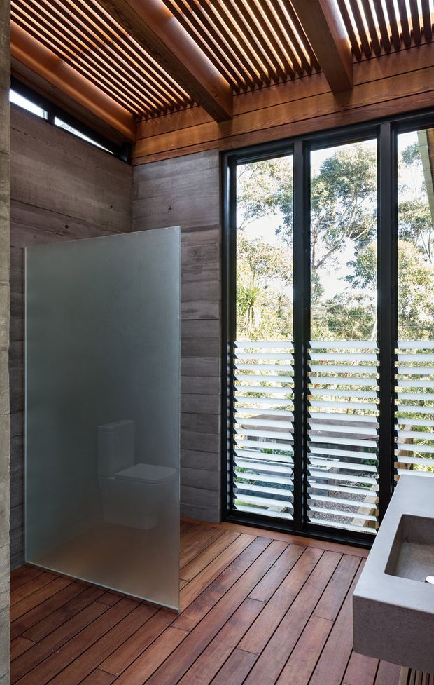 A compact, contemporary bathroom with a frameless frosted-glass shower screen, concrete-look wall panels, dark timber decking-style flooring, and a concrete vanity with integrated basin. Full-height black-framed glazing and horizontal louvres provide strong natural light and views to trees, while exposed timber ceiling battens create a warm architectural feature.