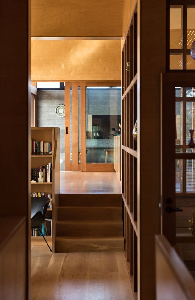 A warm timber-lined interior passage with a short flight of wooden stairs leading to a glazed timber-framed door and a brighter room beyond. Built-in open shelving filled with books lines the left side, while dark timber-framed glazing and joinery occupy the right. The space has strong vertical timber elements, warm ambient lighting, and a compact, crafted residential character.
