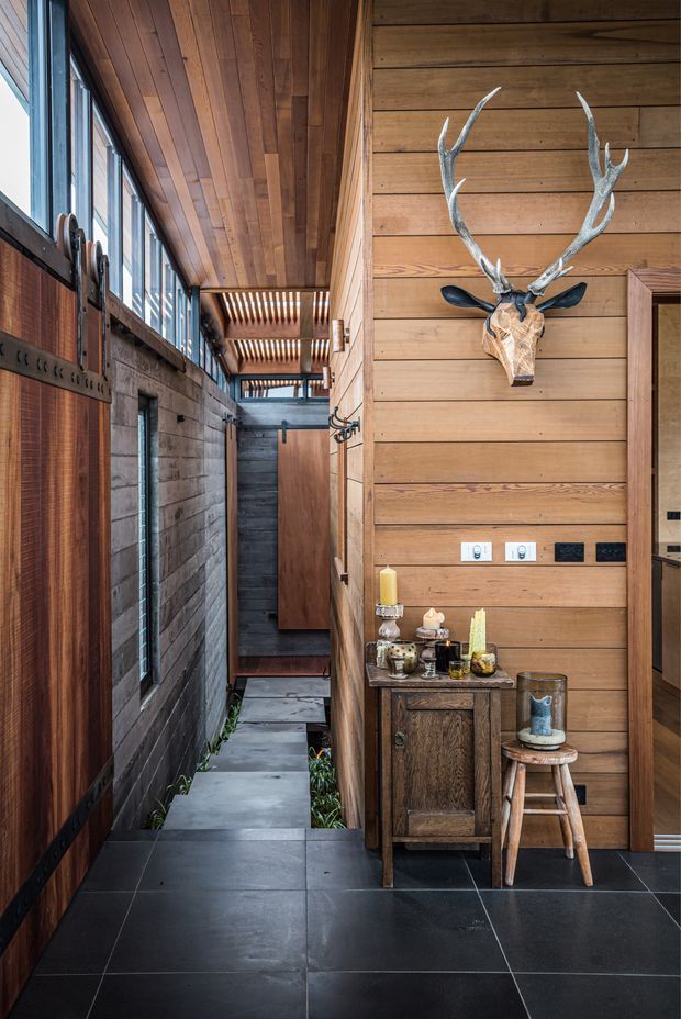 A narrow, warmly detailed interior passage or entry area with dark large-format floor tiles, extensive timber wall and ceiling panelling, tall black-framed glazing along one side, and a small timber console styled with candles and decorative objects. A mounted antlered deer head forms a prominent feature above the console. The passage extends toward a timber door and a partially visible adjoining room.