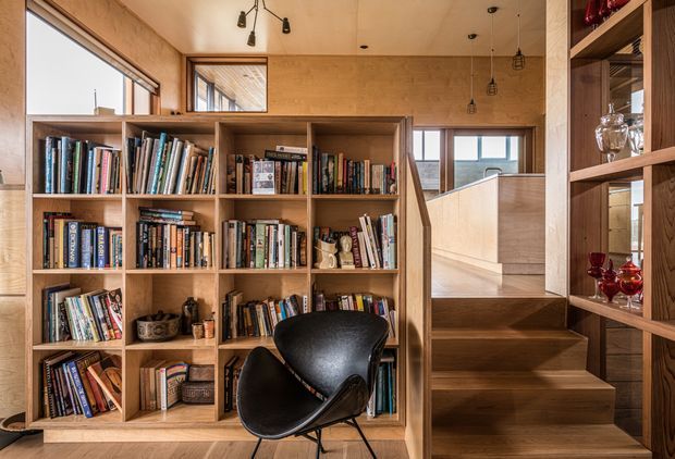 A warm, timber-lined library or reading area with extensive built-in open shelving filled with books. A black leather lounge chair sits in the foreground, while a compact timber stair rises beside the shelving toward a brighter upper level. Natural light enters through high-set windows, and small pendant and track lights provide additional illumination.