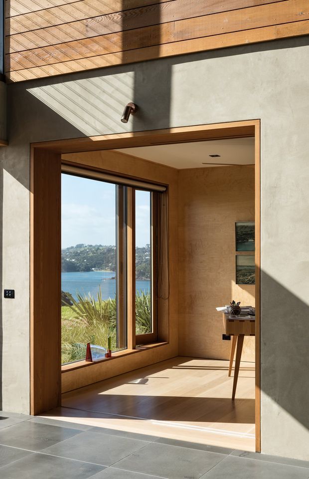 Sunlit architectural interior showing a compact study or workspace opening from a larger tiled area. The room has warm timber-lined walls, ceiling and window reveals, a large timber-framed glazed window overlooking water and a wooded shoreline, a small desk, and a grey textured plaster/concrete wall with a simple wall light. Strong diagonal sunlight and shadows create a calm, refined atmosphere.
