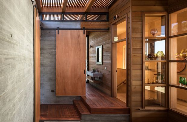 A warmly lit timber-lined interior hallway or entry space with a raised transition and a large sliding timber door. The room features rich dark timber flooring, horizontal timber wall boards, exposed timber slatted ceiling screens, a raw concrete feature wall, built-in glass-fronted display cabinetry, and a small wall-mounted basin visible beyond the door. The composition suggests a refined modern residential entry or circulation space with strong natural-material detailing.