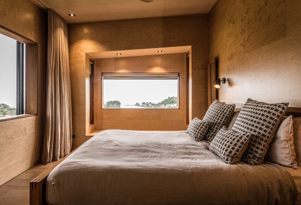 Warm, compact bedroom with a low-profile bed positioned beneath a timber-lined feature wall and framed window. The room has extensive natural timber wall and ceiling finishes, recessed downlights, layered floor-to-ceiling curtains, and patterned neutral bedding. A second window provides views to the landscape, while the overall design feels calm, earthy, and refined.