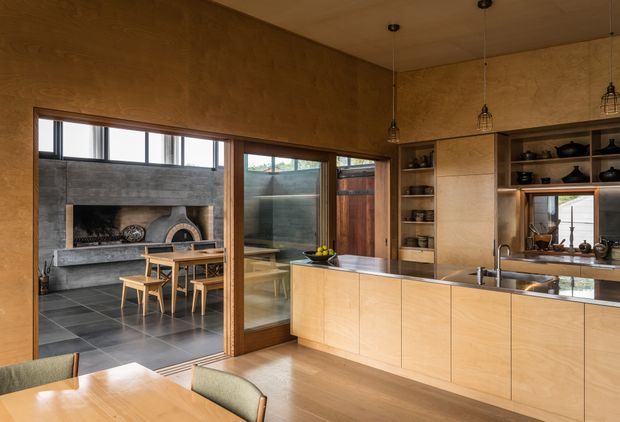 Warm, timber-lined open-plan kitchen and dining area with extensive built-in plywood cabinetry, a large island with integrated sink, open shelving, pendant lights, and a timber dining table. Full-height sliding glazing connects the kitchen to an adjacent outdoor entertaining area featuring a substantial masonry fireplace and dining setting. Dark tiled flooring defines the adjoining space, while the kitchen has a lighter timber floor.