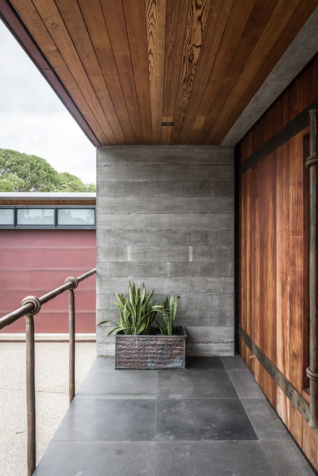A covered exterior entry porch or walkway with dark large-format tiled paving, a richly grained timber soffit, a horizontally textured grey concrete wall, and a large timber pivot door with dark metal detailing. A metal handrail borders the walkway, with a planter of architectural foliage at the end. A pink-red rendered building wing is visible across the open side.