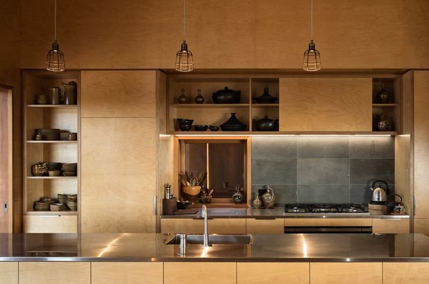 A warm, compact contemporary kitchen with extensive light timber veneer cabinetry, open display shelving, a long island bench with sink, and integrated cooking appliances. The backsplash is formed from large dark grey metal or stone panels, while three black wire pendant lights hang above. Dark ceramics and cookware are displayed in the open shelving, creating a refined, earthy atmosphere.