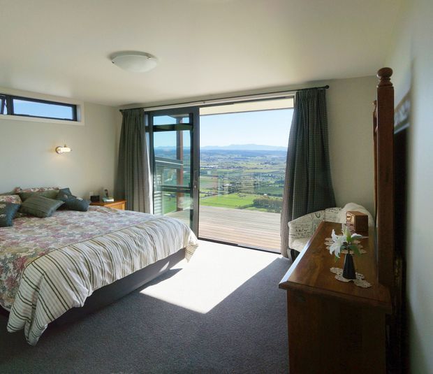 A bright bedroom with a double bed, floral and striped bedding, grey carpet, patterned curtains, and a timber dresser. Large glazed doors open onto a balcony or deck with expansive rural valley views, while a high horizontal window and wall light provide additional daylight.