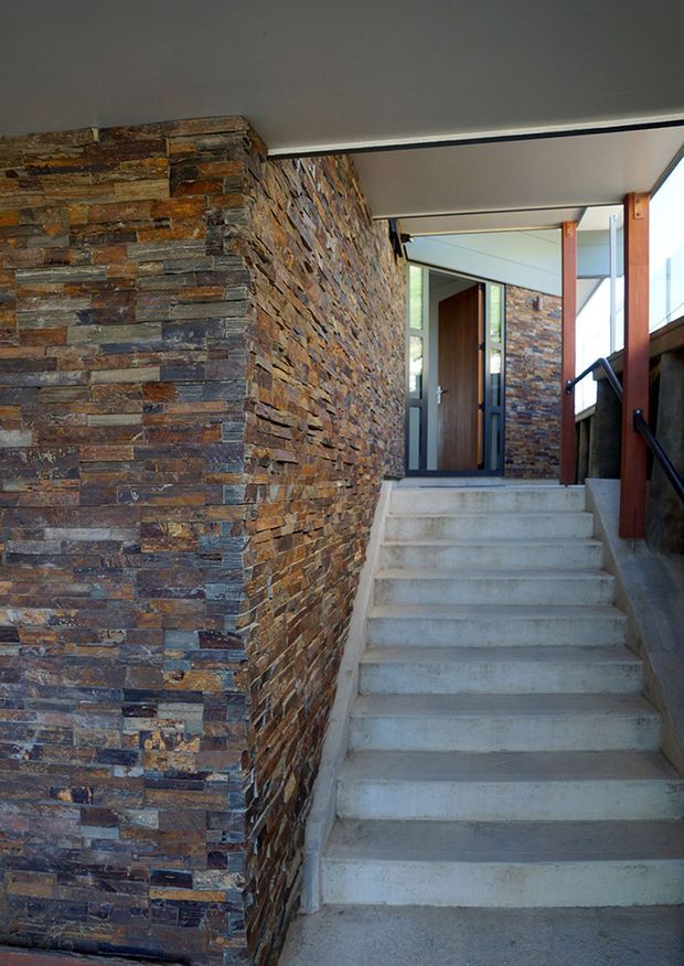 A covered exterior entry stairway rises between substantial stacked-stone walls toward a modern timber and glass entrance door. The stairs are raw concrete, with dark metal handrails, timber posts, and a flat soffit overhead. The stonework has varied brown, grey, rust, and slate tones, creating a rugged contemporary architectural entrance.