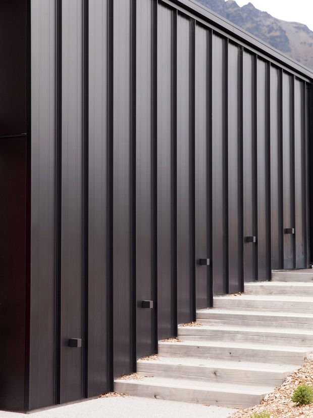 Exterior architectural detail showing a long dark vertically ribbed metal-clad facade beside a stepped concrete walkway. Repeated narrow vertical panels and small dark fixtures create a strong rhythmic composition, with rugged mountain terrain faintly visible in the background.