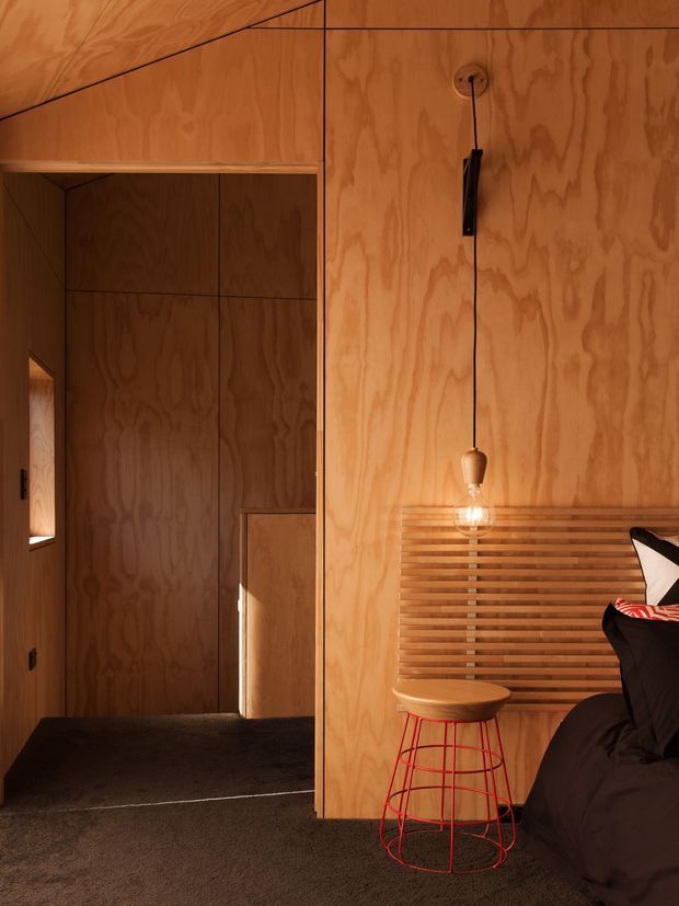 Warm, compact bedroom interior clad extensively in natural plywood, with a dark carpeted floor, integrated plywood wardrobe or hallway joinery, and a timber slatted feature headboard. A hanging pendant light, wooden stool with red wire frame, and dark bedding add contrast. The image has a highly integrated, minimalist timber-lined design with strong geometric panel joints and warm ambient lighting.