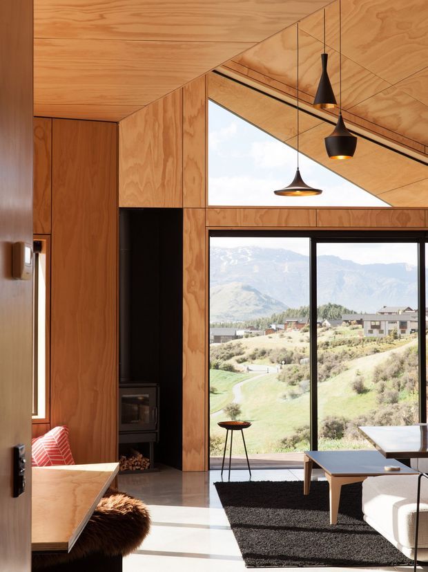 A contemporary open-plan living and dining space lined with warm plywood panels and cabinetry. A dramatic sloped timber ceiling incorporates a high triangular window, while full-height glazing frames a broad mountain and rural landscape. Black pendant lights, a dark fireplace recess, low-profile furniture, and a textured black rug create a warm, refined interior with strong natural light.