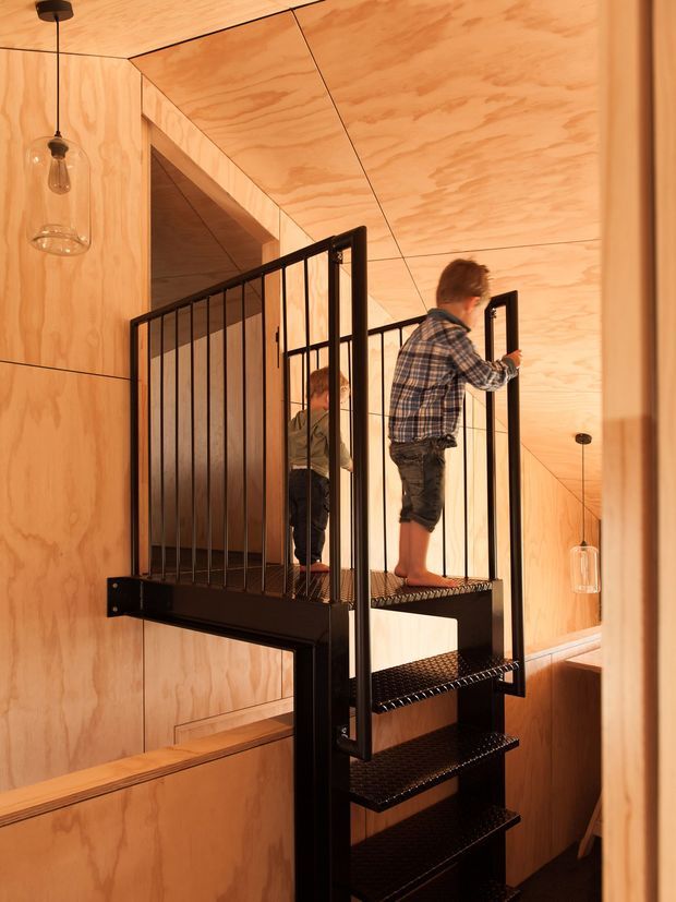 Interior view of a compact modern stairway and landing clad in warm plywood panels. A black metal open-riser staircase with grated treads and slender vertical balustrades rises through the space, with children on the stairs. Exposed plywood-lined walls and ceiling create a cohesive timber-lined interior, while simple pendant lights provide warm illumination.