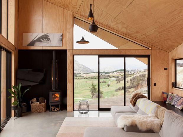 Warm contemporary living room in a compact timber-lined dwelling, featuring plywood walls and vaulted ceiling, expansive black-framed glazing with mountain and rural views, a black wood-burning stove, pendant lights, and a pale sectional sofa. The room has a refined natural palette and strong indoor-outdoor connection.