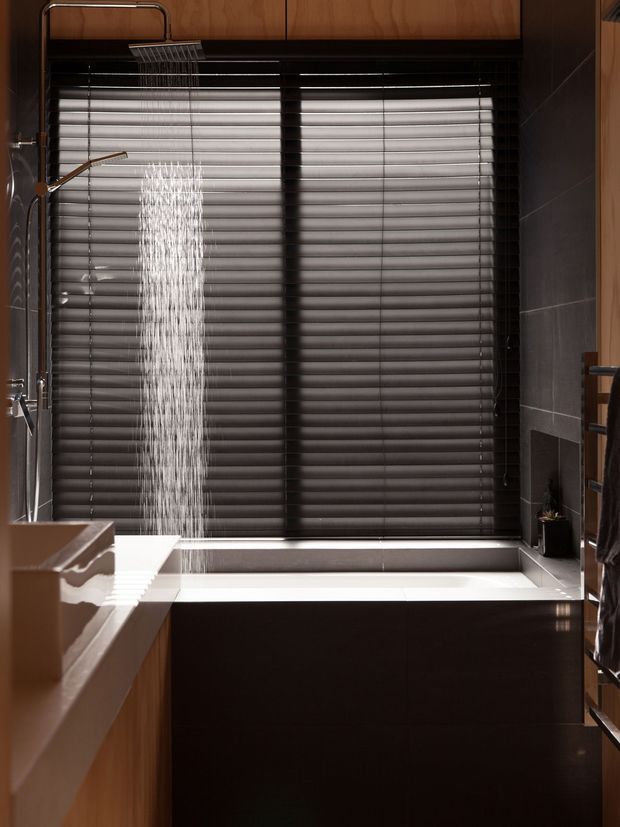 A compact, dark-toned bathroom featuring a freestanding rectangular bathtub positioned beneath a large window with dark horizontal blinds. A ceiling-mounted rain shower falls beside the tub, with a separate wall-mounted shower fitting visible at left. The room has dark grey tiled walls, glossy dark timber cabinetry or panelling, warm timber overhead cabinetry, and a white tub rim and countertop edge. The composition is vertical and tightly framed, with a refined, moody contemporary aesthetic.