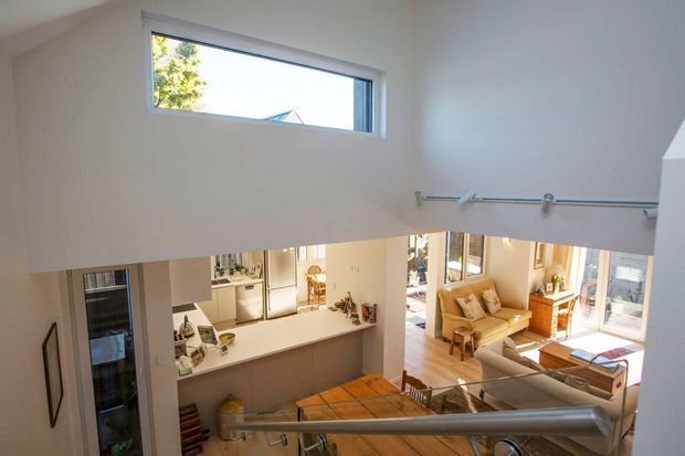 View from an upper-level stair landing into a bright, open-plan home. A timber stair handrail and metal balustrade overlook a kitchen with an island or peninsula, dining area, and sunlit living room with large windows and glazed doors. The space has white walls, timber flooring, and a high open stairwell with a narrow horizontal window.