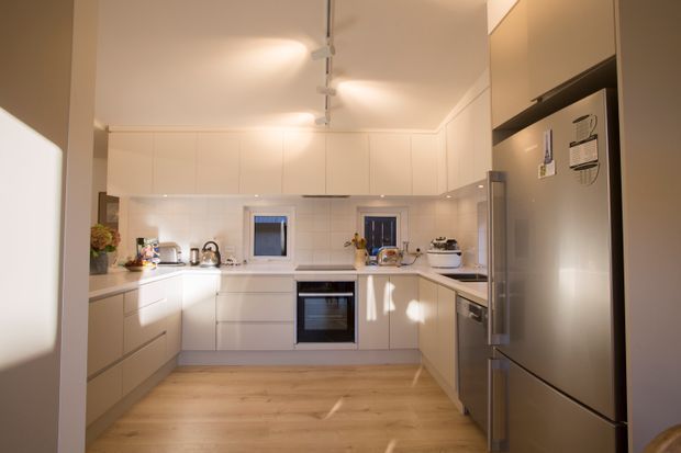 A bright, compact contemporary kitchen with white handleless cabinetry arranged in a U-shaped layout, pale engineered-looking benchtops, stainless-steel appliances, and light timber-look flooring. Track lighting runs along the ceiling, while windows and a glazed door bring in natural light.