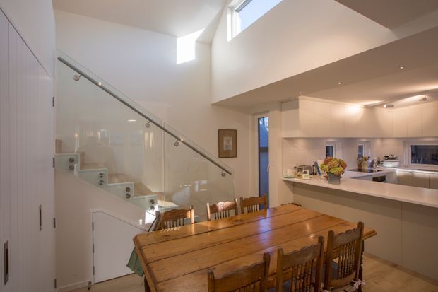 Bright open-plan dining and kitchen area with a large rustic timber dining table and matching chairs in the foreground. A contemporary white kitchen with a long island bench occupies the right side, while a glazed staircase with a simple metal handrail rises to the left. High-level windows and a partial void bring in natural light, creating an airy, warm space.