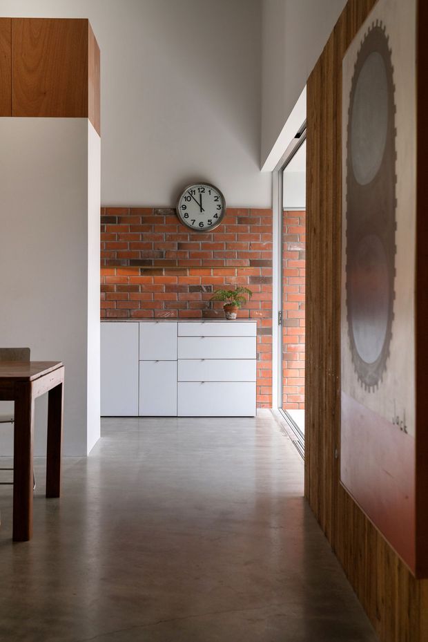 A minimalist interior hallway or open-plan living area with polished concrete flooring, white walls, exposed red brickwork, and built-in white cabinetry. Warm timber wall panelling and a large framed artwork line the right side, while a timber dining table is partially visible on the left. A large window or glazed opening brings in natural light, and a wall clock and small potted plant provide simple accents.