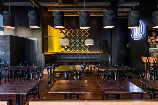 A dark, atmospheric commercial dining venue with rows of timber tables and black metal chairs, a long upholstered banquette, exposed ceiling services, and oversized ribbed pendant lights. Black walls and joinery contrast with warm timber flooring, a vivid yellow feature wall, and a geometric patterned screen behind the seating. A bar or service area is visible at the right.