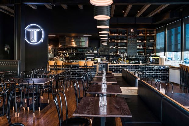 A stylish dark-toned bar and restaurant interior with numerous timber dining tables, black bentwood chairs, upholstered booth seating, a long tiled bar counter, extensive bottle shelving, pendant lights, and exposed dark ceiling beams. A neon logo sign and large windows add visual highlights.