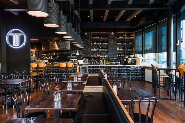A dark, industrial-style restaurant or bar interior with rows of timber dining tables, black bentwood chairs, upholstered banquette seating, and a long tiled bar. The space features black-painted ceilings with exposed beams, hanging ribbed pendant lights, large windows, extensive bottle shelving, and a backlit logo sign.