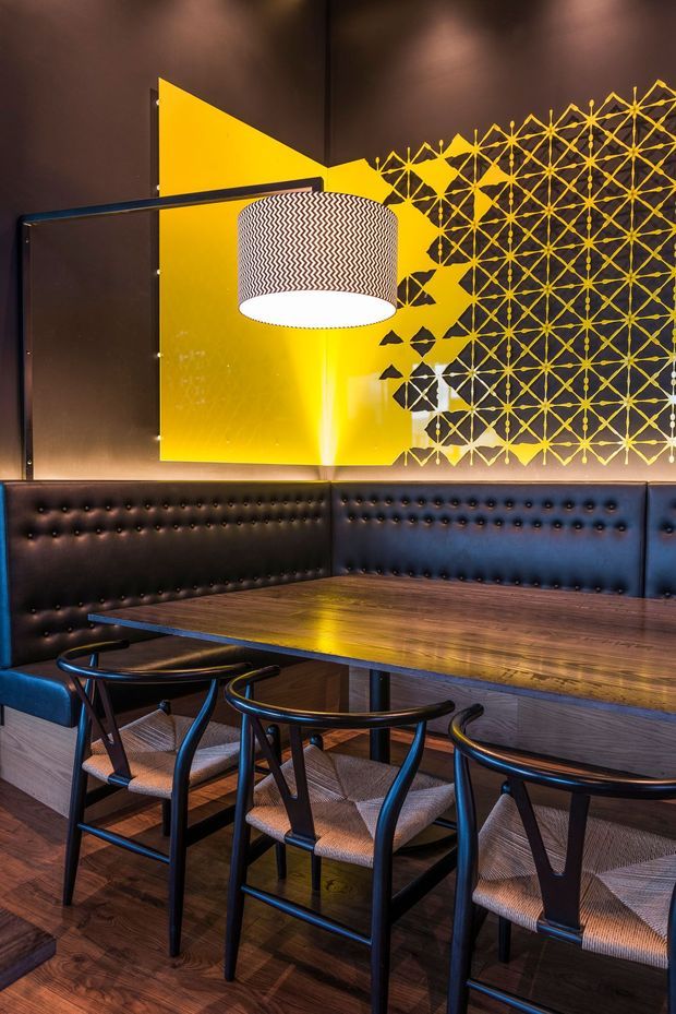 A stylish restaurant or hospitality dining booth with a long dark timber table, black tufted banquette seating wrapping around the corner, and several black wood-framed chairs. A large patterned pendant light hangs over the table. The walls feature dark finishes with a bold yellow geometric mural or screen, creating a dramatic, graphic interior.