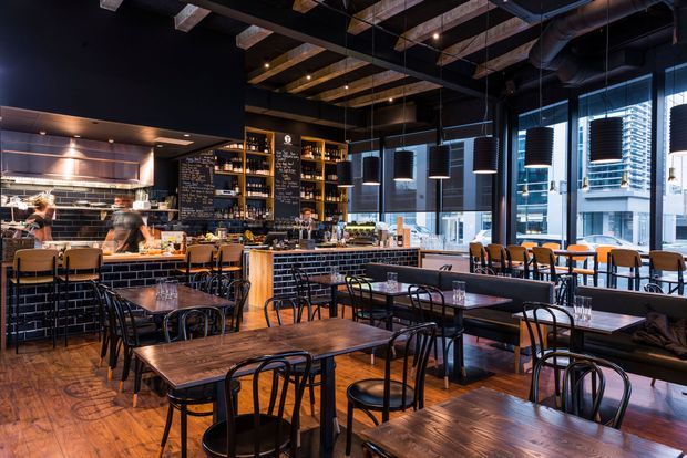 A dark, industrial-style restaurant or café interior with numerous timber dining tables and black metal chairs. The space features a long bar and open kitchen, exposed timber ceiling beams, visible mechanical services, black tiled walls, pendant lighting, chalkboard menus, and full-height windows overlooking an urban setting.