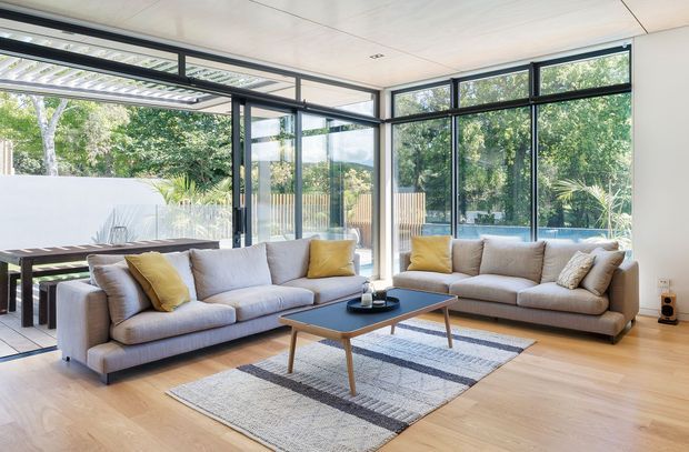 Bright contemporary living room with two large neutral sofas, mustard accent cushions, a low timber and black coffee table, and a geometric area rug. Full-height black-framed glazing opens the room visually to a covered outdoor dining area and leafy garden, while light timber flooring and a white panelled ceiling create an airy, relaxed interior.