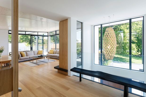 Bright, open-plan contemporary living area with extensive black-framed glazing, pale timber flooring, and a built-in timber-lined structural partition. A light upholstered sectional sofa, coffee table, dining area, and long black bench are visible, with a landscaped garden beyond the windows and a large woven pendant feature outside or adjacent to the glazing.
