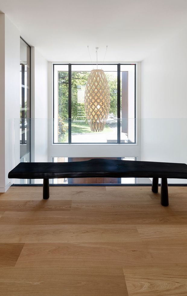 Bright minimalist interior featuring a long sculptural black timber bench on a light oak floor. Full-height glazing and a large projecting window overlook greenery, while a woven wood pendant light hangs centrally in the double-height space. White walls and slim dark window frames create a clean, contemporary composition.