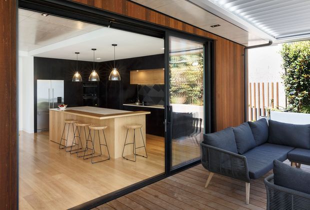 Contemporary open-plan kitchen opening onto a timber deck and outdoor lounge through large black-framed sliding glass doors. The kitchen features a pale timber island with seating, dark cabinetry, pendant lighting, timber flooring, and a timber-lined exterior wall and ceiling over the deck.