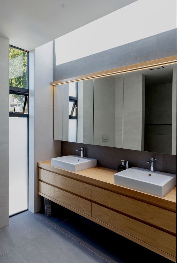 Contemporary bathroom with a long double vanity, two white vessel basins, wall-mounted mirrored cabinetry, warm timber drawers, dark grey wall surfaces, and large grey floor tiles. Tall glazing brings in abundant natural light.