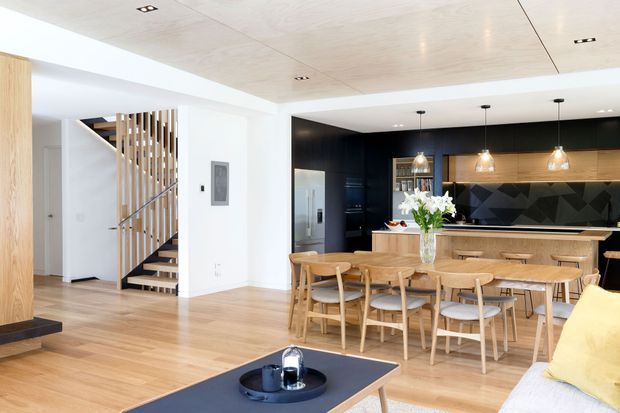 Bright open-plan kitchen and dining area with a large timber island, six timber dining chairs, dark cabinetry, geometric dark splashback, pendant lights, and integrated stainless-steel appliances. The room opens to a living area and features a timber-slatted staircase, light timber flooring, and a pale timber ceiling.