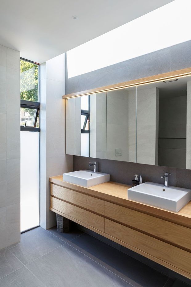 Bright contemporary bathroom featuring a long double vanity with white vessel basins, natural timber drawers, a dark grey tiled backsplash, large mirrored wall cabinets, grey floor and wall tiles, and a high clerestory-style window providing abundant natural light.